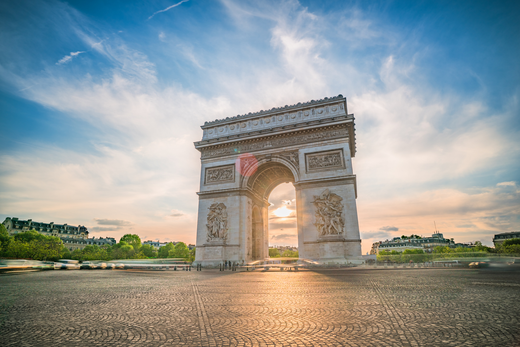 A photo of Paris' famed Arc de Triomphe, a massive arch of carved white stone in the middle of a traffic circle, seen at sunset.