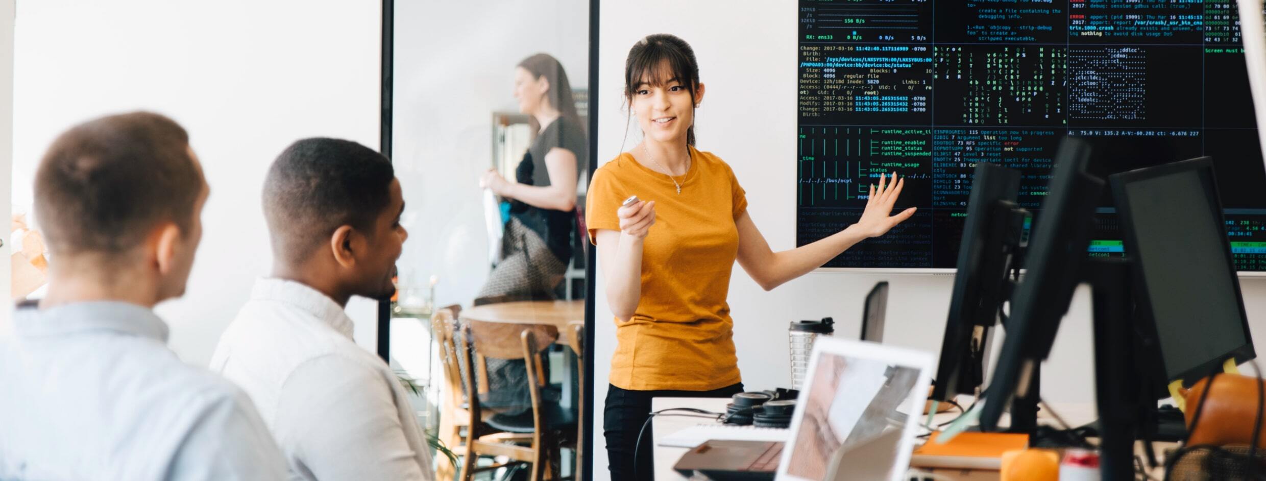 stock photo of a woman standing at a large screen displaying code while leading a small meeting